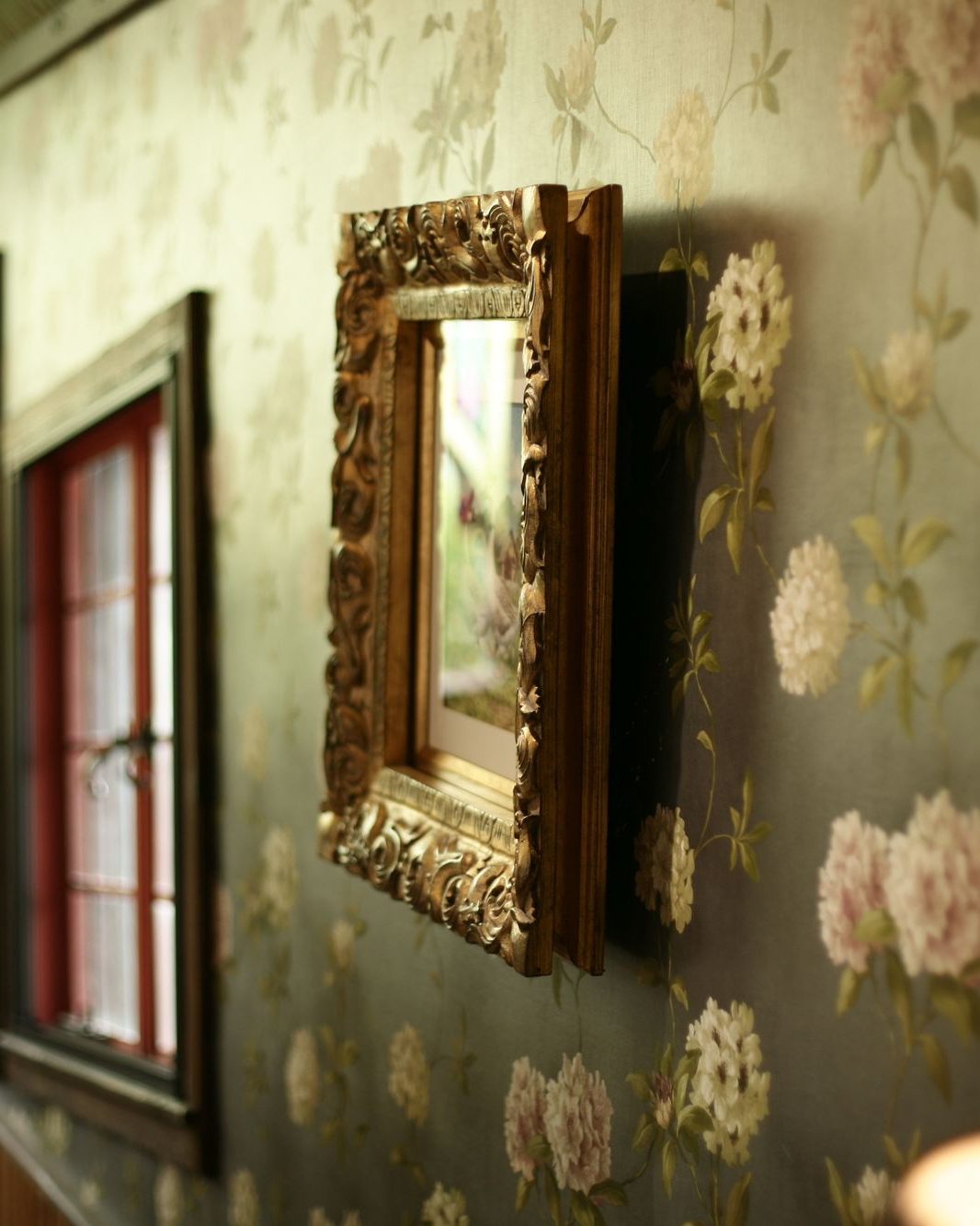 Traditional sage-toned floral wallpaper with a gilded carved mirror frame near a window