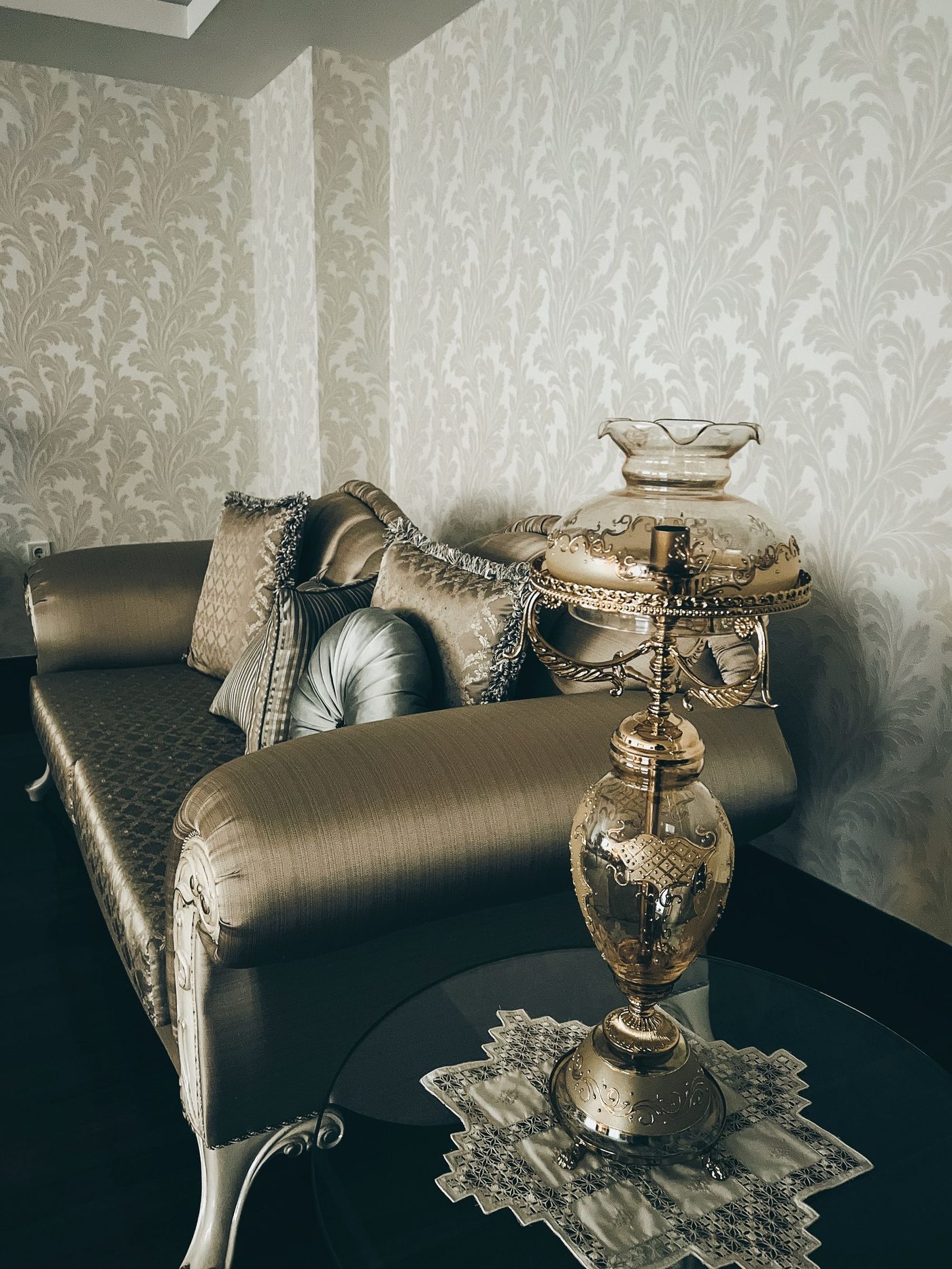 Formal sitting room with an ornate gold acanthus-leaf damask wallpaper wall behind a tailored sofa and crystal table lamp